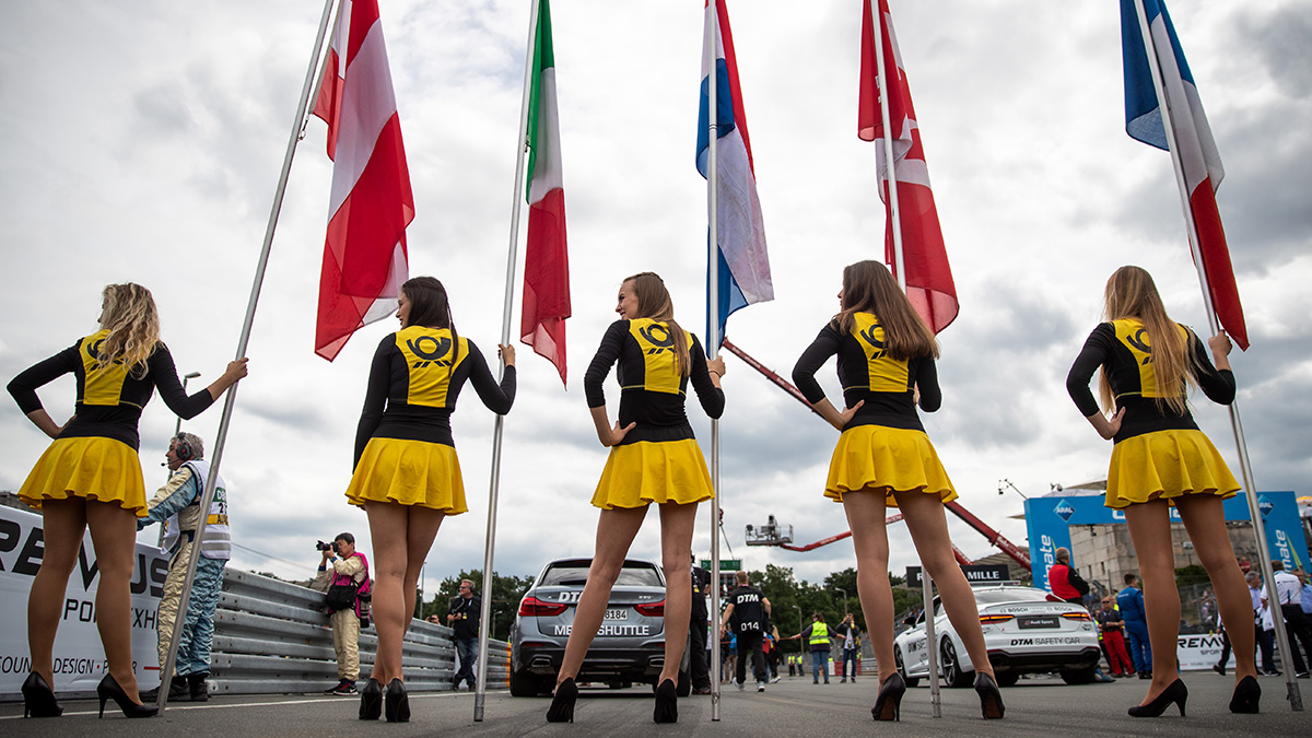 Five girls in short black and yellow dresses stand in a line holding flags from different countries at the start of a car race