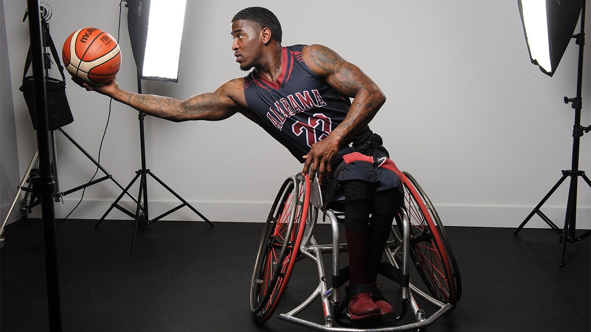 Dequel Robinson in University of Alabama basketball jersey sitting in wheelchair with outstretched arm holding basketball