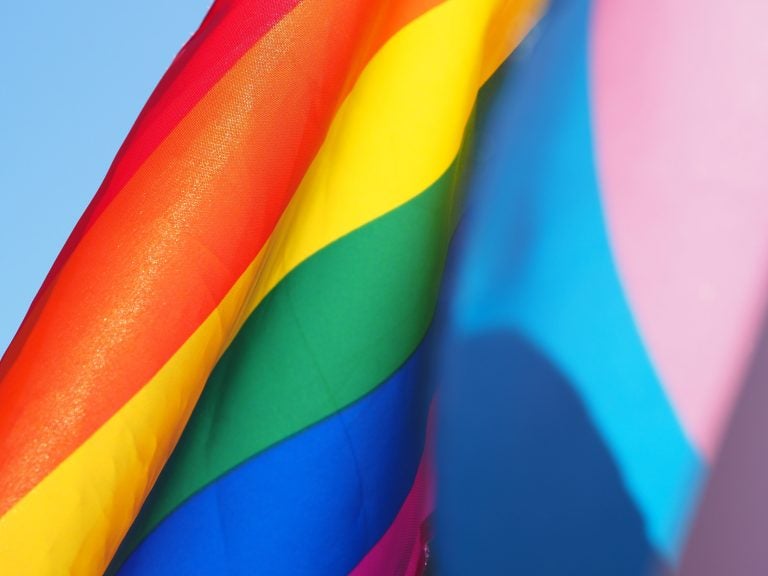 A rainbow colored flag against a blue sky, with a pink and blue flag close up in front of it. (Photo by Cecilie Johnsen on Unsplash)