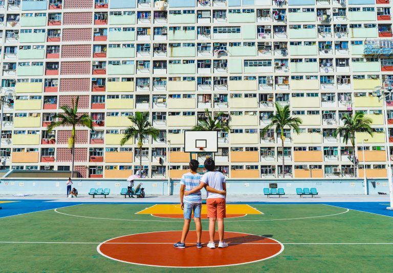 Two young children stand arm in arm on a colorful basketball court with a colorful housing building in the background